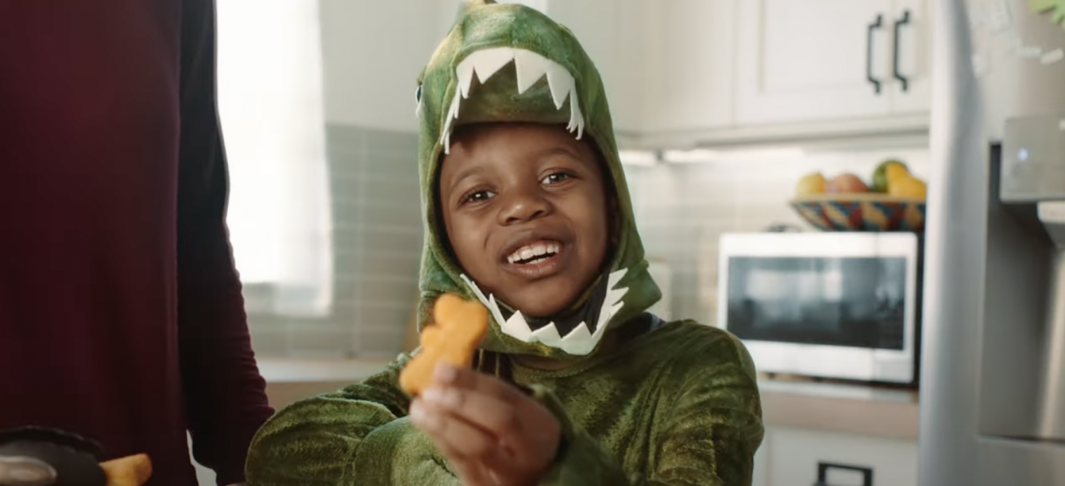 Corn Kid wears a dinosaur costume and holds up a Dino Veggie Tot