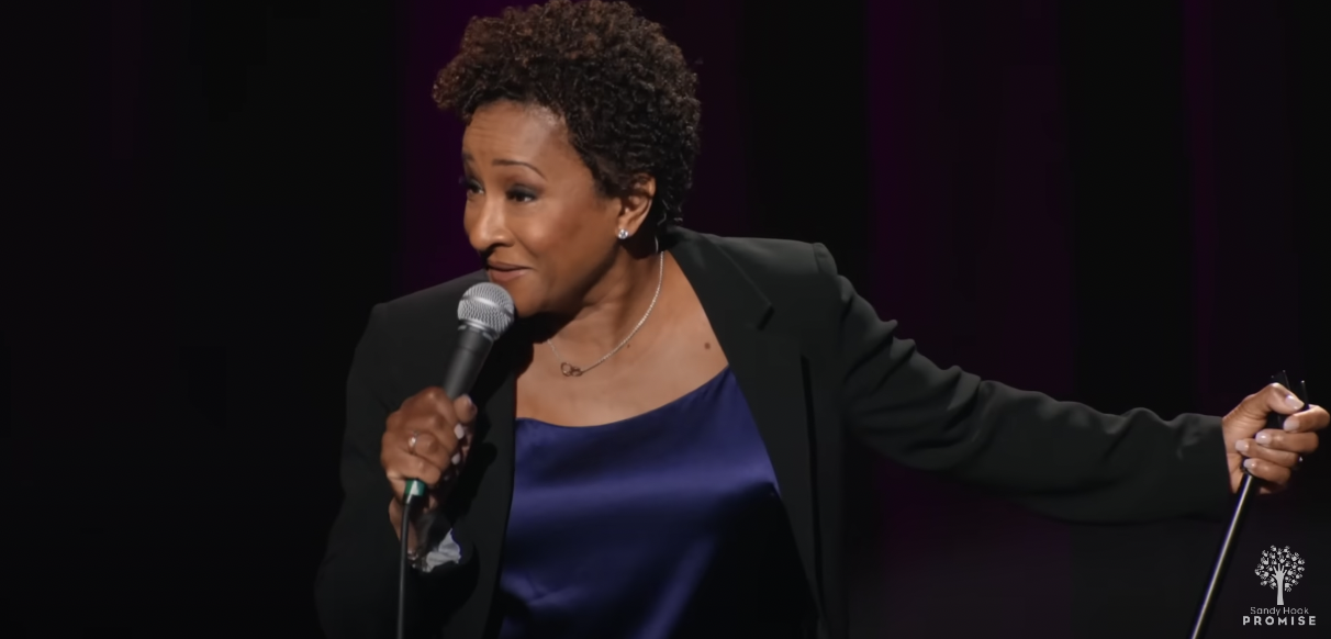Wanda Sykes wears a blue blouse and a black blazer and holds a microphone