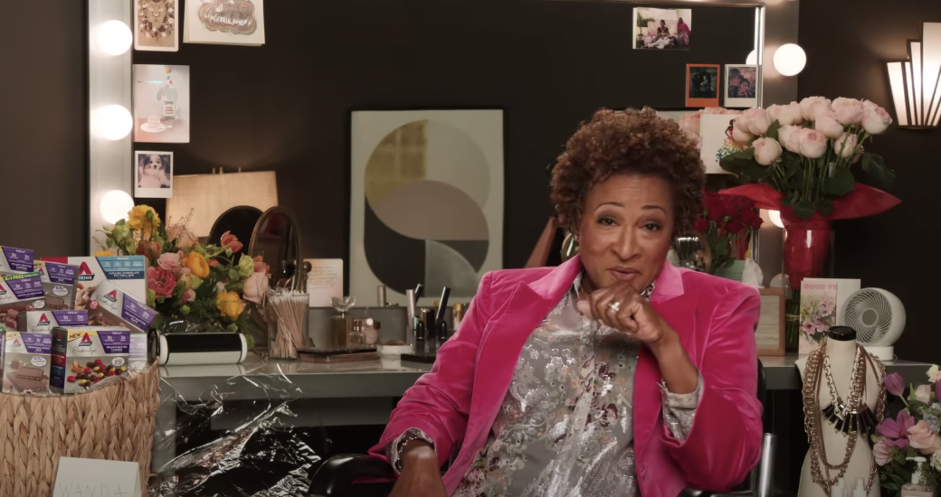 Wanda Sykes wears a pink blazer in her dressing room adorned with photographs and a candy basket