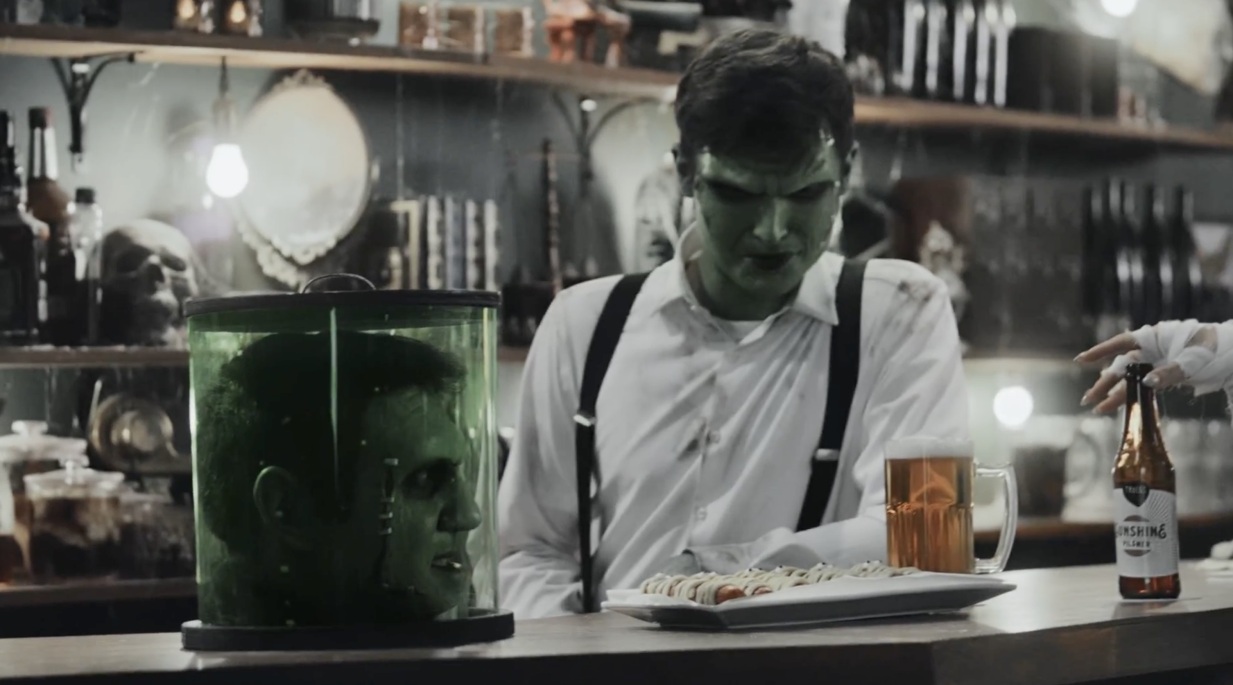 A Frankenstein monster head in a jar rests on a pub bar. Behind the bar is another Frankenstein monster.