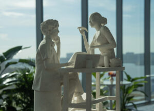Two figures carved in stone; one sits before a laptop on a desk, the other sites atop the desk eating a slice of pizza