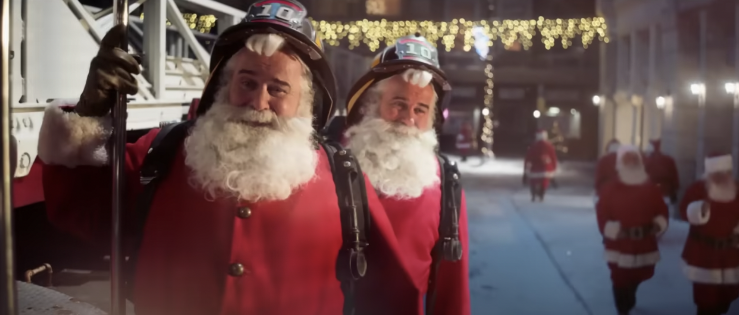 Two Santas ride a firetruck down a street populated with other Santas