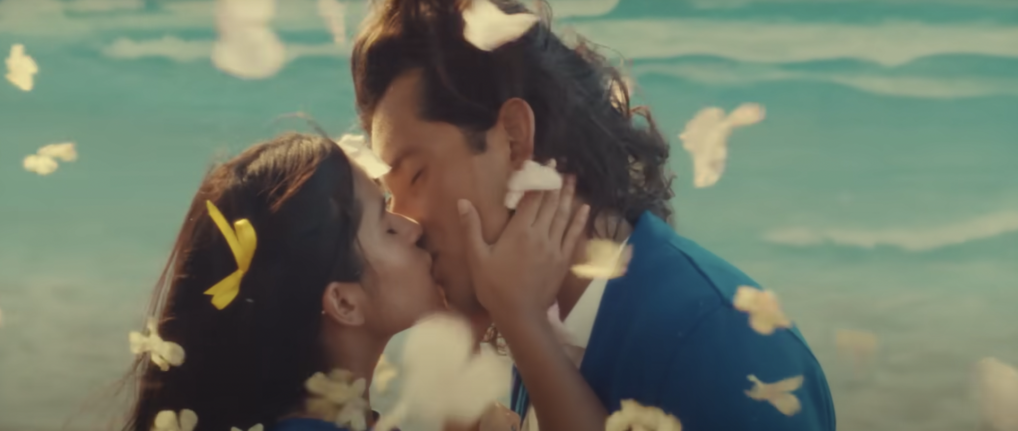 Two young people kisss with petals falling around them in front of a beach backdrop
