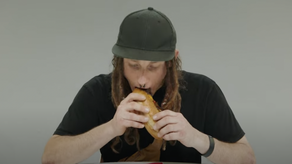 A person wearing a black shirt and hat bites into a sandwich
