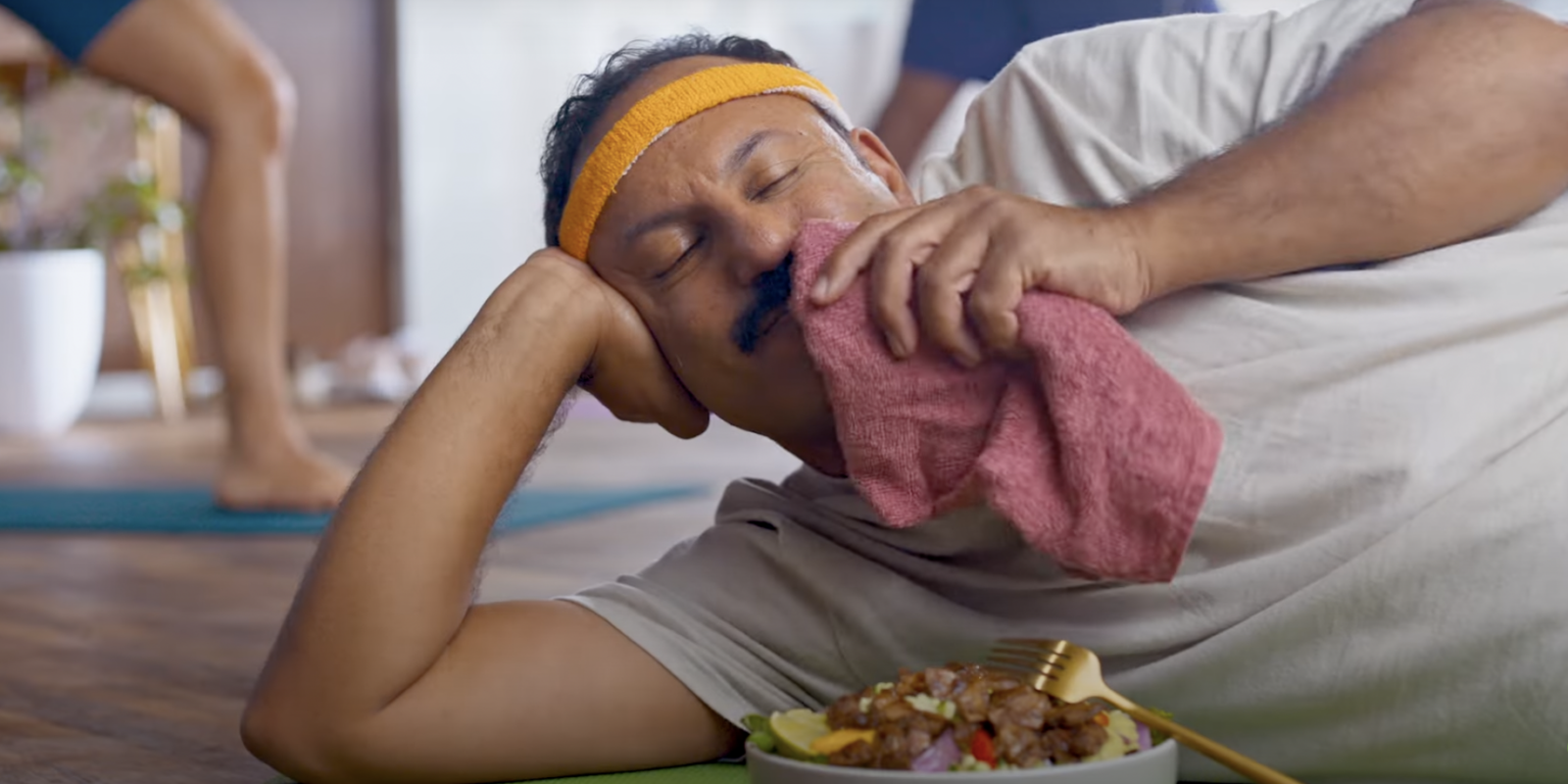 A man wearing a sweat headband dabs his mouth with a napkin while lying on his side eating a bowl of Beyond Steak.