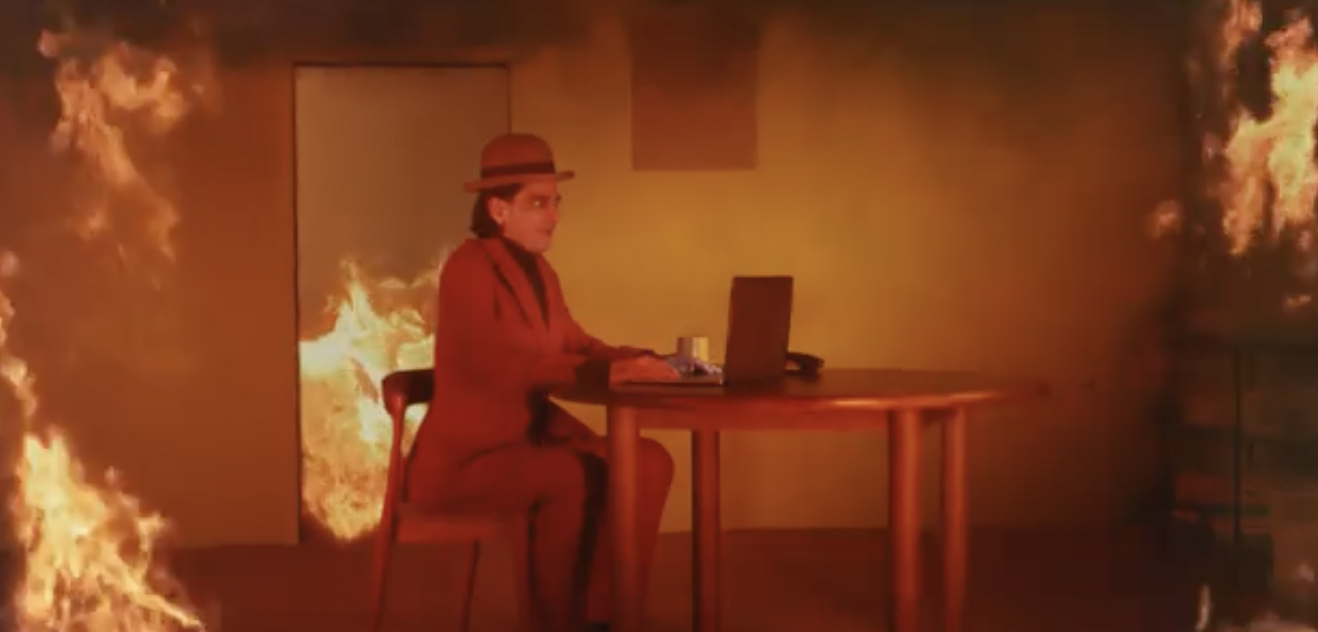 A live recreation of the This is Fine meme with a man in a hat seated at a table while the room is on fire