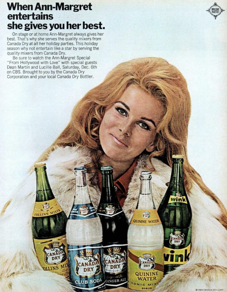 A woman wearing a fur coat and holding glass bottles of Canada Dry products. "When Ann-Margret entertains she gives you her best."