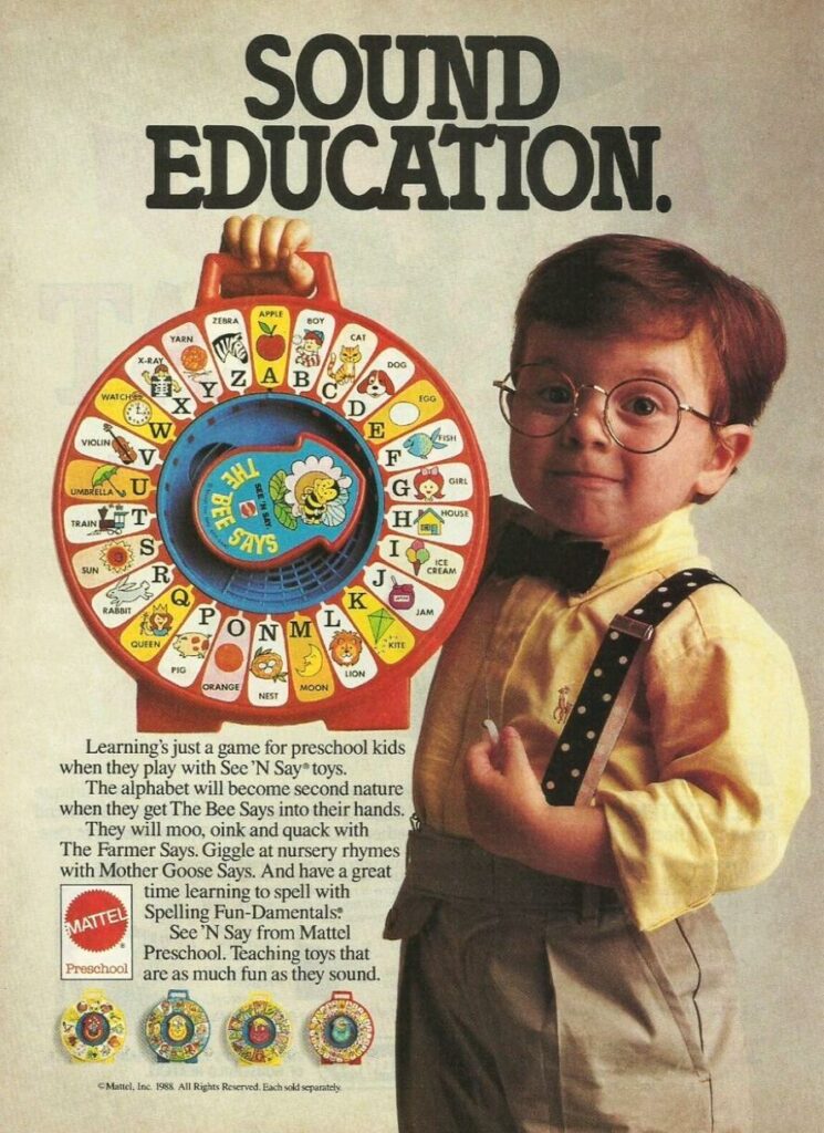 A little boy wearing glasses and suspenders holds up a See 'n Say toy