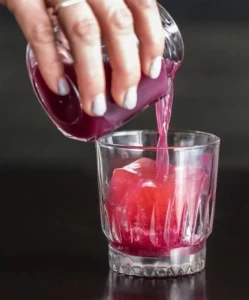 A hand pours a glass jar of grape juice into a cocktail glass