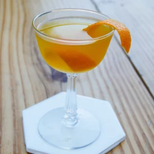 An orange cocktail in a coupe glass on an octagonal coaster garnished with orange twist